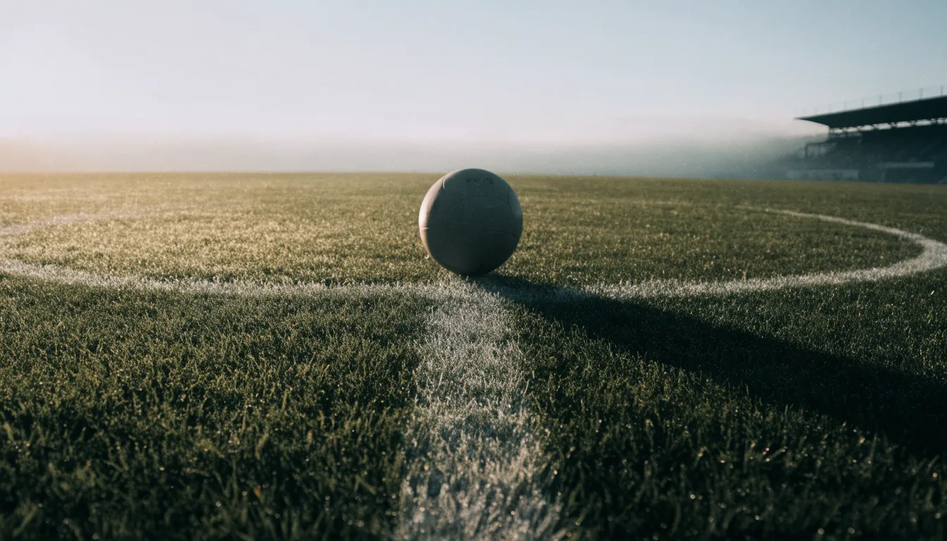 Ein Fußball liegt auf dem Mittelkreis eines leeren Rasenplatzes am frühen Morgen, im Hintergrund eine ferne Tribüne im Dunst.
