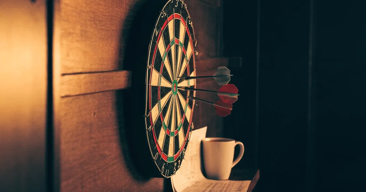 Eine Dartscheibe an einer Holzwand in einem schwach beleuchteten Raum, drei Pfeile stecken nahe der Mitte, warmes Licht von der Seite.